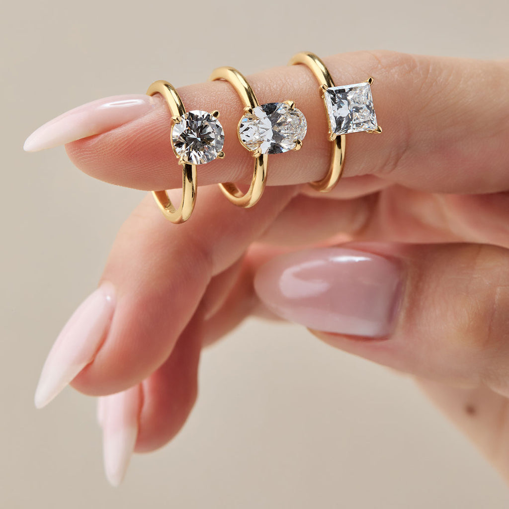 Tres anillos de compromiso de oro amarillo con solitarios de diamante o moissanita, llevados en un dedo. Los anillos tienen diferentes cortes: redondo brillante, ovalado y princesa cuadrado. Anillos de pedida de oro minimalistas y clásicos.
