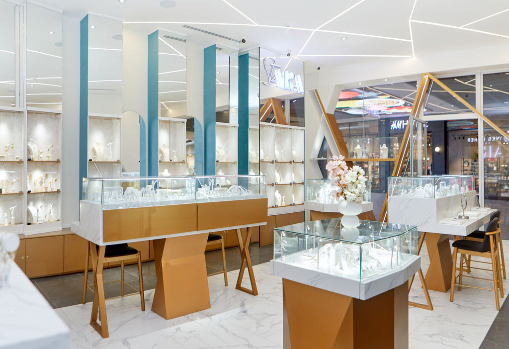 Interior de la joyería ARA en Oakland Place, Guatemala. El diseño de la tienda es moderno con decoración blanca y dorada, suelos de mármol blanco y detalles en azul. Las vitrinas de cristal exhiben la colección de joyería, incluyendo collares, anillos y aretes. El nombre de la marca ARA es visible en un letrero de neón.