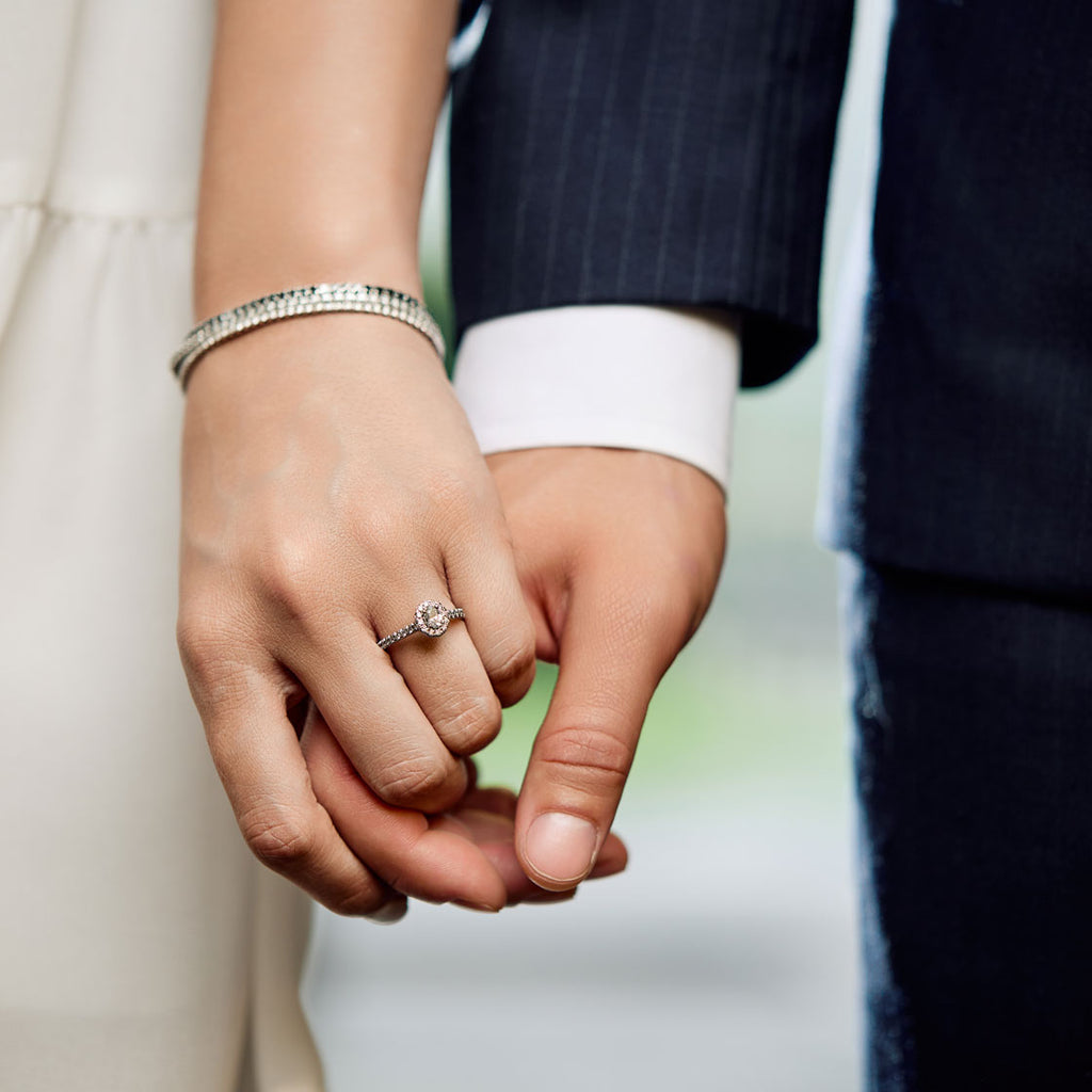 Primer plano de una pareja tomándose de la mano, mostrando un anillo de compromiso de la colección ARA Weddings. El anillo de compromiso es un diseño halo con un diamante o gema rosa central y pavé en la banda. La mujer también lleva una pulsera tipo brazalete de diamantes. Anillos de pedida y alianzas de matrimonio.