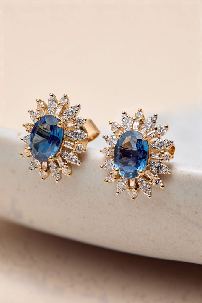 Par de aretes stud o de botón de oro amarillo con diseño tipo sol o halo. Cada arete tiene una piedra preciosa central ovalada de color zafiro azul, rodeada por un halo de diamantes baguette y redondos. Pendientes de alta joyería con zafiros y diamantes para boda o evento especial.