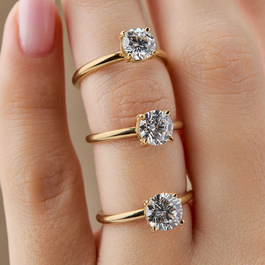 Tres anillos de compromiso solitarios de oro amarillo que ilustran las diferencias en el peso en quilates (Carat) del diamante. Los anillos muestran diamantes redondos en un engaste de cuatro puntas, con variaciones sutiles de tamaño. Anillos de pedida con diamantes de 0.75ct, 1.00ct y 1.25ct para comparación.