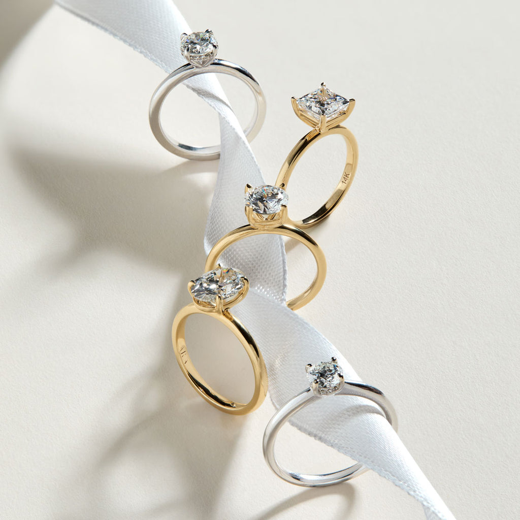 Cinco anillos de compromiso solitarios de diamante en diferentes cortes y metales, sobre una cinta blanca. La selección incluye anillos de oro amarillo y oro blanco con diamantes en corte redondo, ovalado y princesa cuadrado. Muestra de diferentes engastes de anillos de pedida.