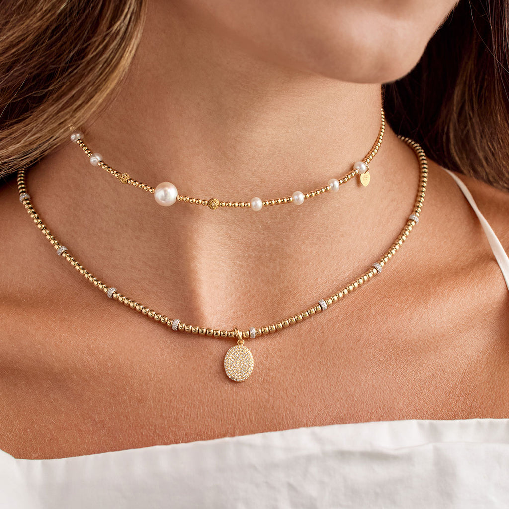 Layering de collares de oro y perlas. La imagen muestra dos chokers de joyería apilable: un collar superior de cuentas de oro con perlas de río y dijes de corazón, y un collar inferior de cuentas de oro con un dije redondo pavé con circonitas o diamantes. Collares finos de oro para mujer.